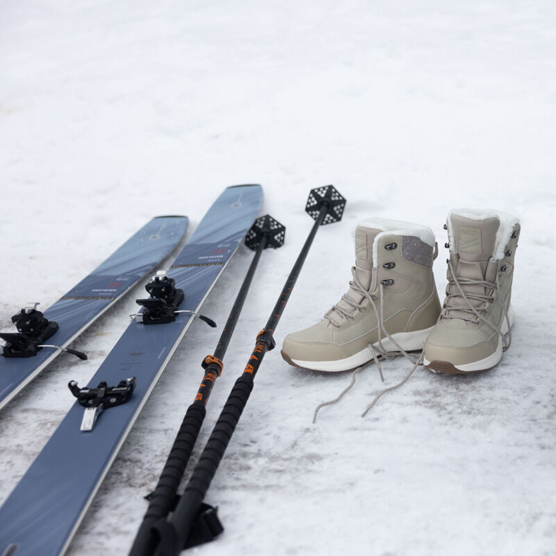 Ett par skidor, skidstavar och Halti Dundee Mid DX Vinterskor Dam placeras på snön.