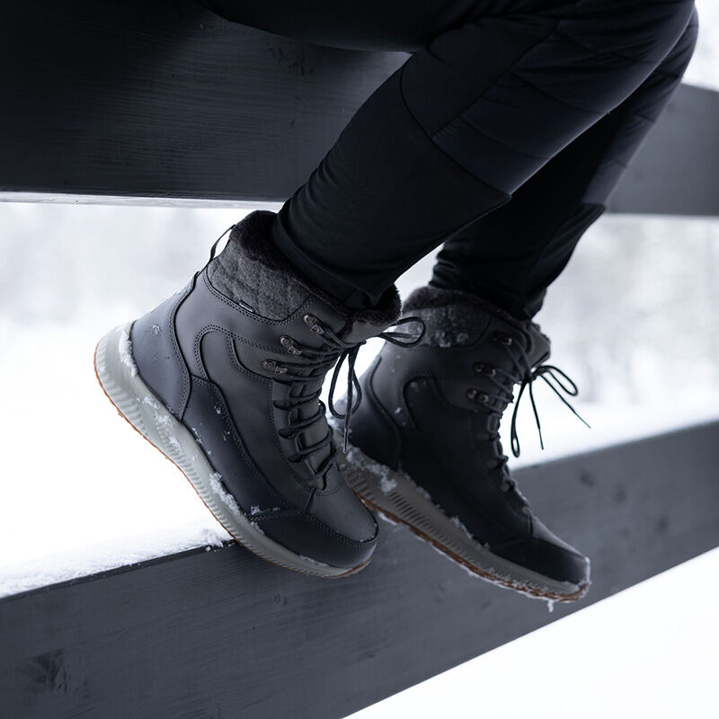 En person bär Halti Dundee Mid DX Vinterskor Dam och svarta byxor, sittande på ett snötäckt staket utomhus.