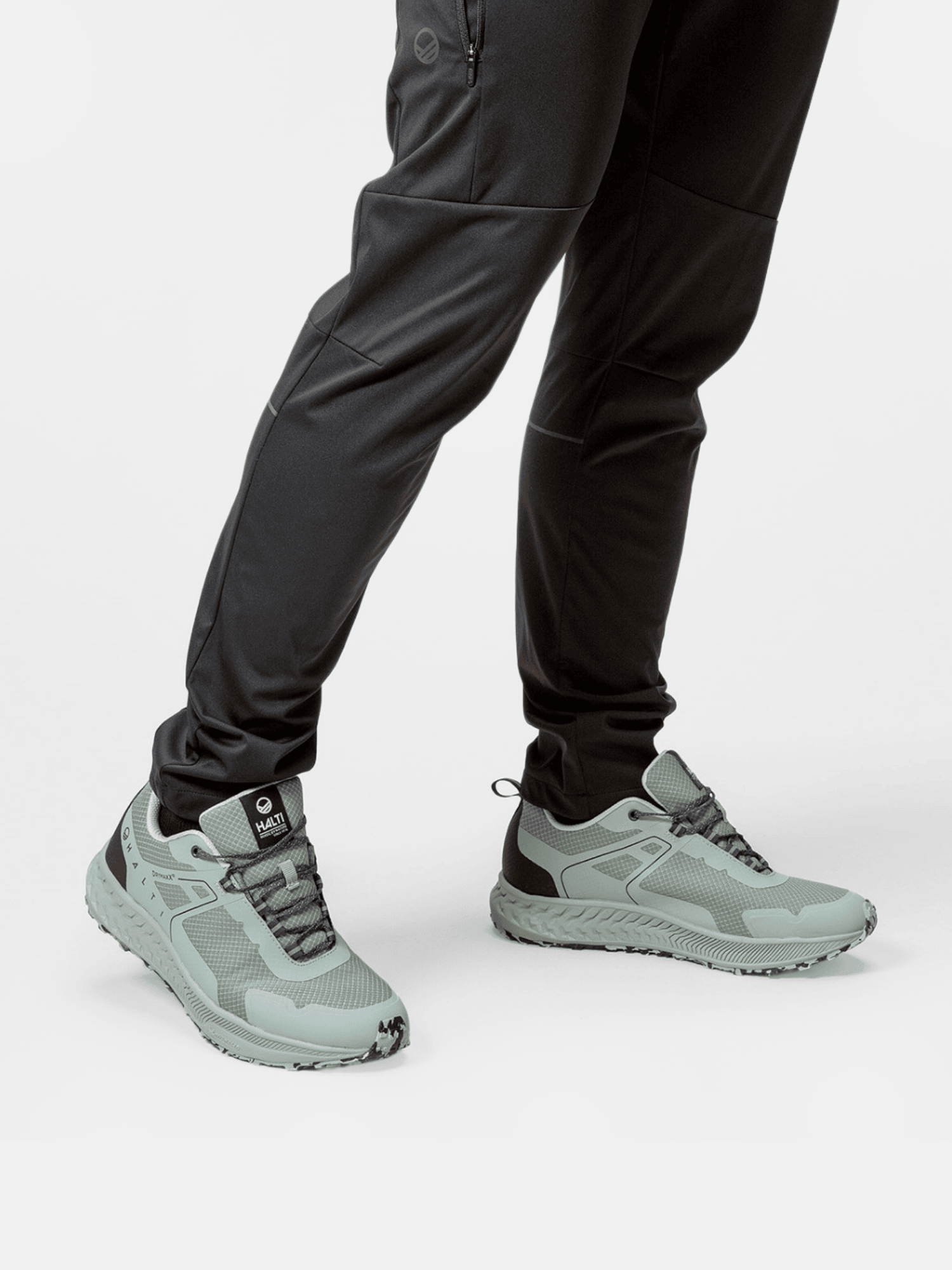 Person i svarta joggingbyxor och Halti Pallas Low 2 DrymaxX Hybrid Sneaker Herr, stående mot en vit bakgrund.