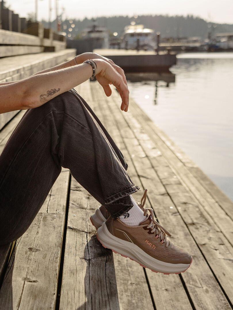 En person i svarta jeans och Halti Rapid Sneaker Dam sitter på träplankor vid vattnet med båtar i bakgrunden.