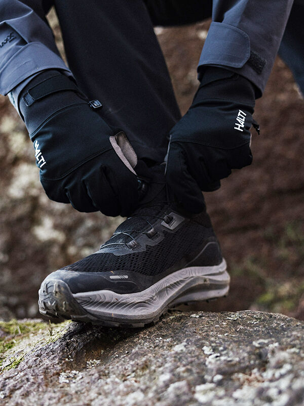 En person i svarta handskar knyter Halti Rapid Mid Freelock DX Skor Unisex snören på en sten under stiglöpning.