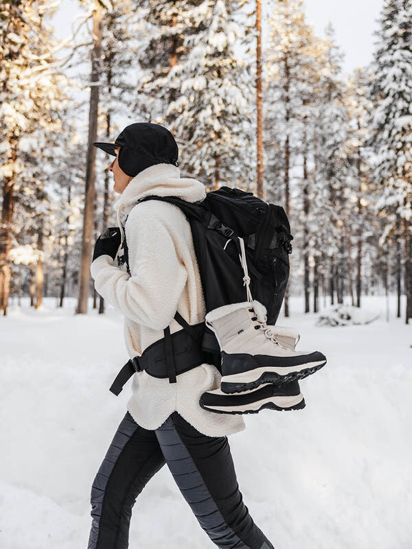 Person i Halti Tornio 2 DX vinterskor bär svart ryggsäck, går genom snöig skog.
