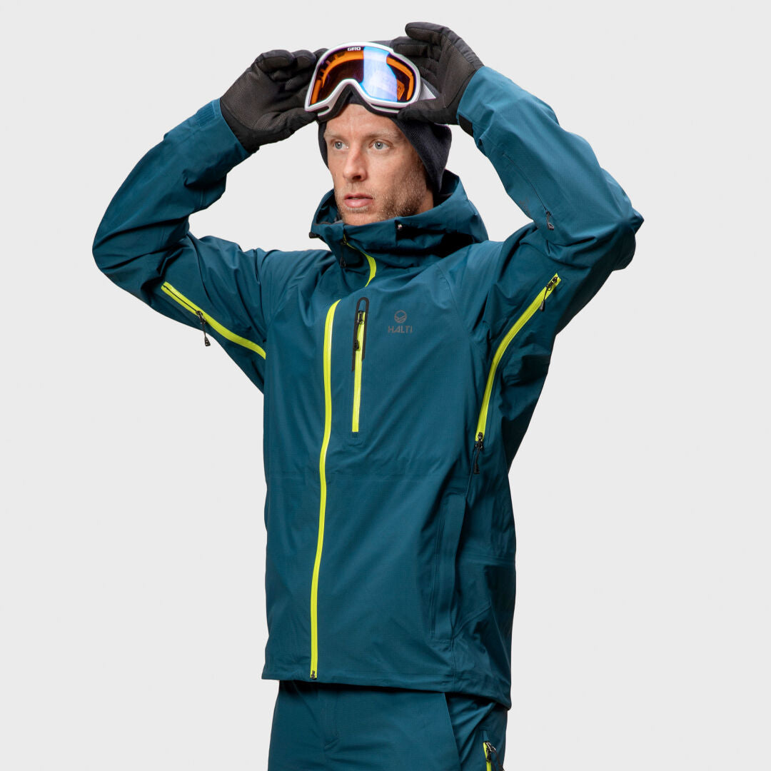 Någon i en blågrön Halti Alpine Unisex 3L DrymaxX Skaljacka tar på sig skidglasögon mot en enfärgad ljus bakgrund.