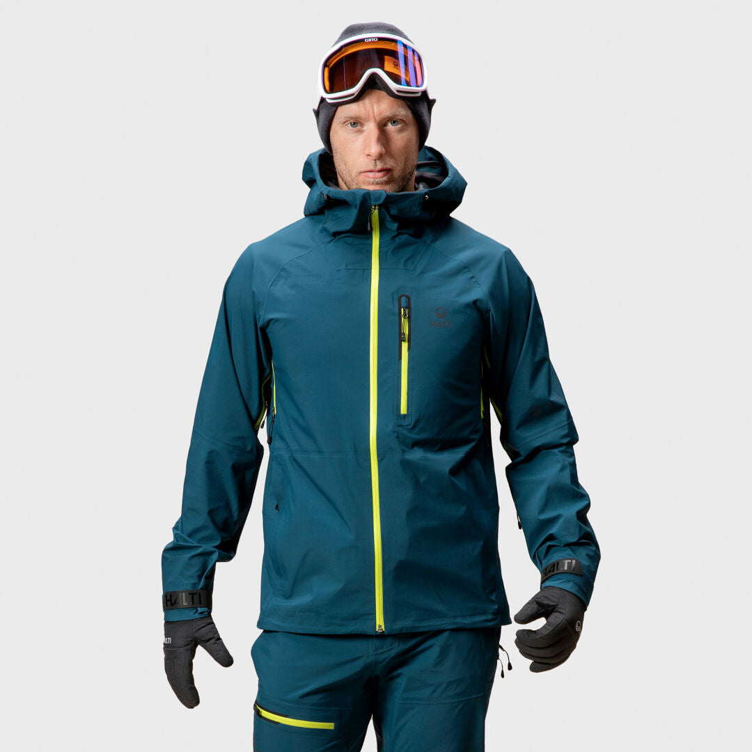 En man i en blå Halti Alpine Unisex 3L DrymaxX skaljacka och skidglasögon står vänd framåt mot en slät bakgrund.