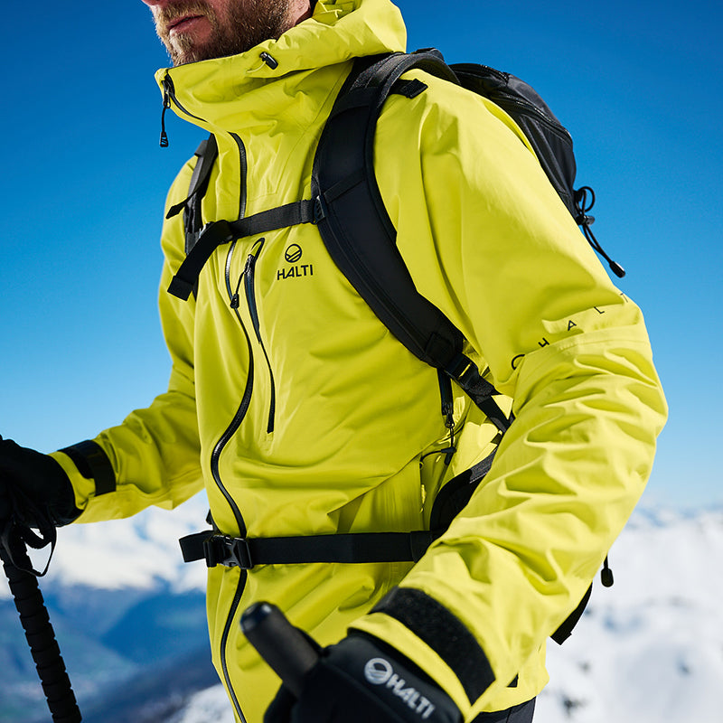 Person i ljusgul Halti-jacka och handskar håller i skidstavar och står utomhus med snöiga berg i bakgrunden.