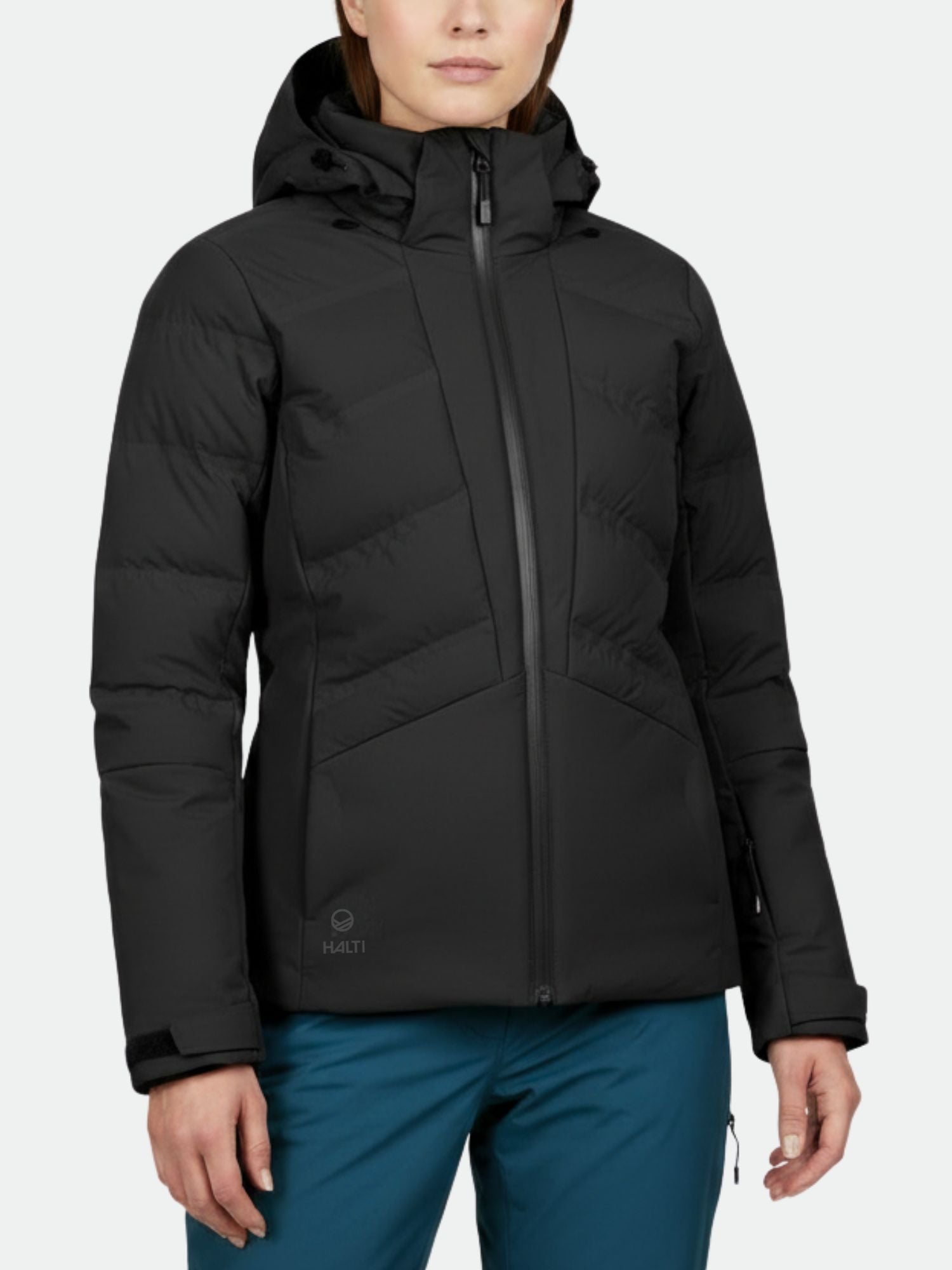 En person bär en svart Halti Nordic Arcty Skidjacka Dam med dragkedja framtill och blå byxor, mot en enfärgad bakgrund.