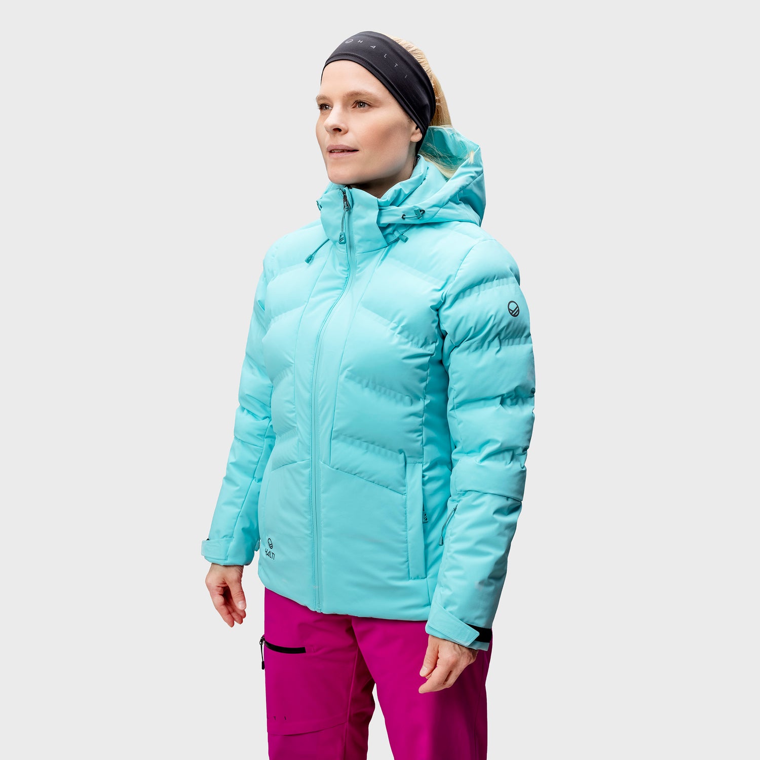En kvinna i ljusblå pufferjacka, rosa byxor och svart pannband står mot en enfärgad bakgrund.