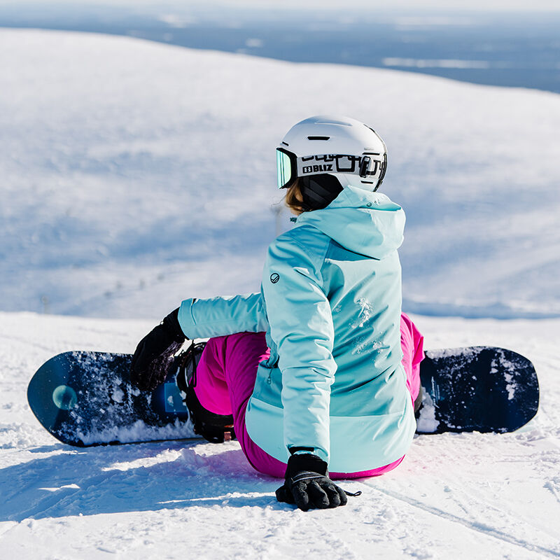 En person i en Halti Wedeln DrymaxX Skidjacka och rosa byxor sitter på snö med snowboard, hjälm och goggles.