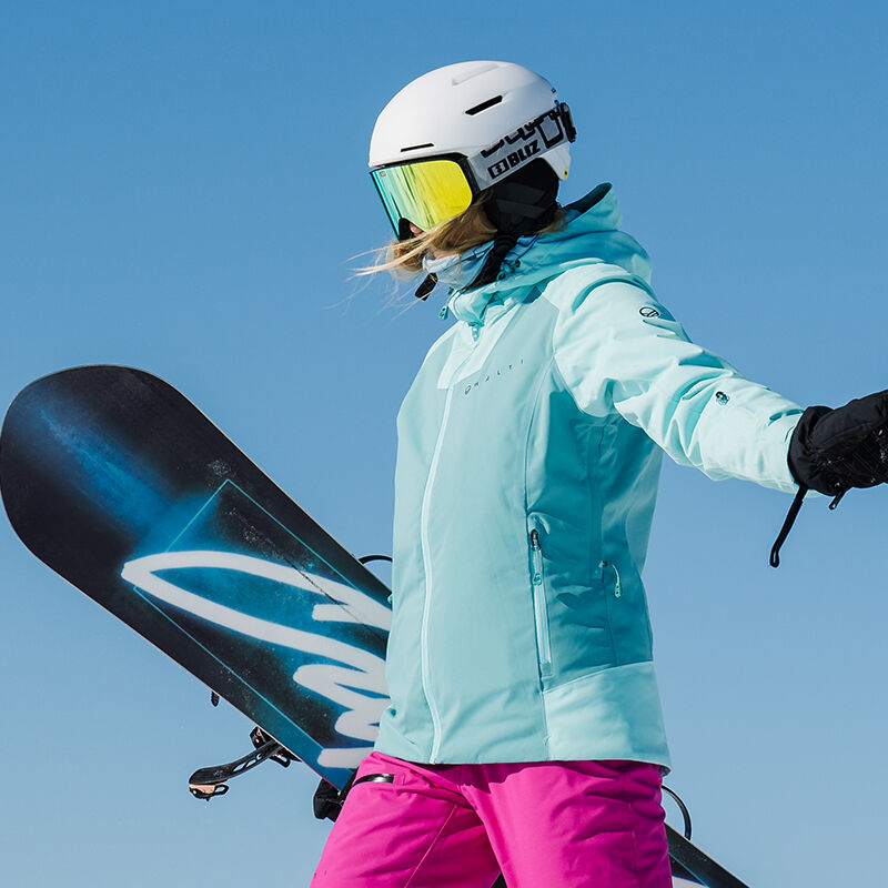 Kvinna i Halti Wedeln DrymaxX skidjacka håller snowboard mot klarblå himmel, klädd i rosa byxor och hjälm.