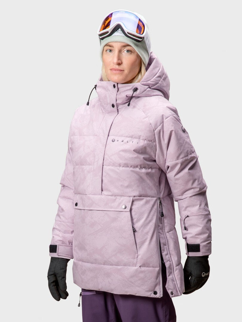 En person i ljuslila Halti Chowper Skidanorak Dam, skyddsglasögon, handskar och grå mössa står mot en enfärgad bakgrund.