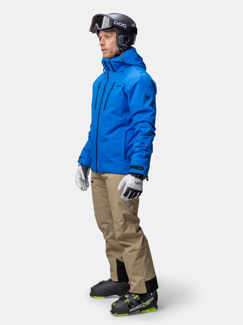 En man i Halti Vertica DX Skidjacka Herr, solbruna byxor, pjäxor, handskar och hjälm med skyddsglasögon står upprätt.