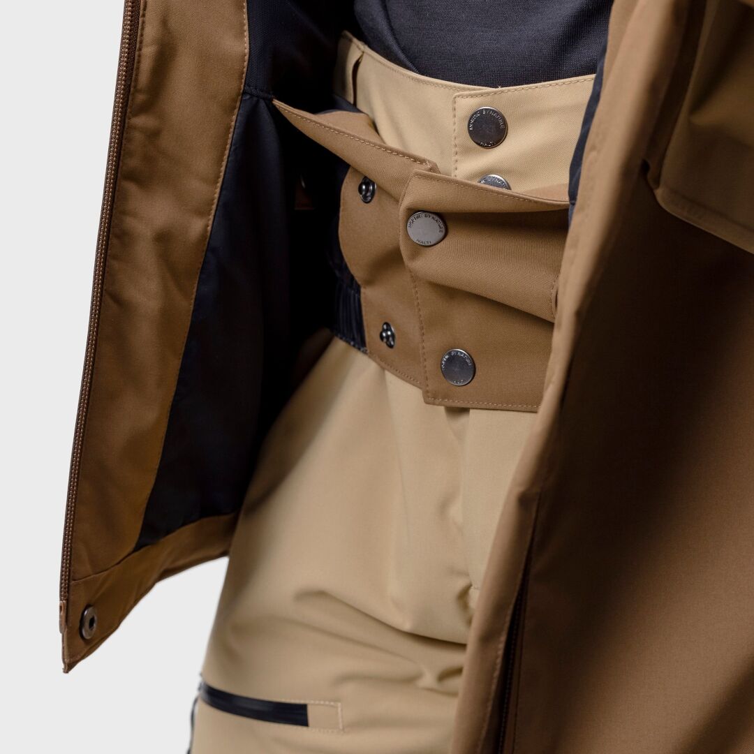 Närbild på en person i beige byxor med hög midja, mörk skjorta och Halti Planker DX Skidjacka Herr av återvunnet material.