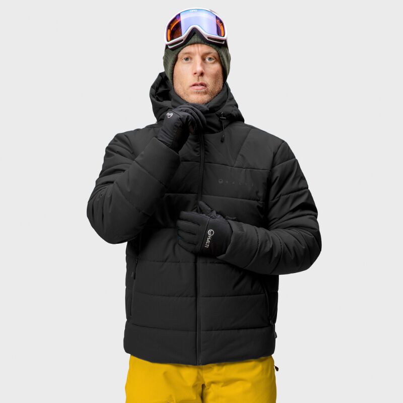 En person i en Halti Mellow Puffer Skidjacka Herr, gula byxor och skidglasögon står mot en enfärgad ljus bakgrund.
