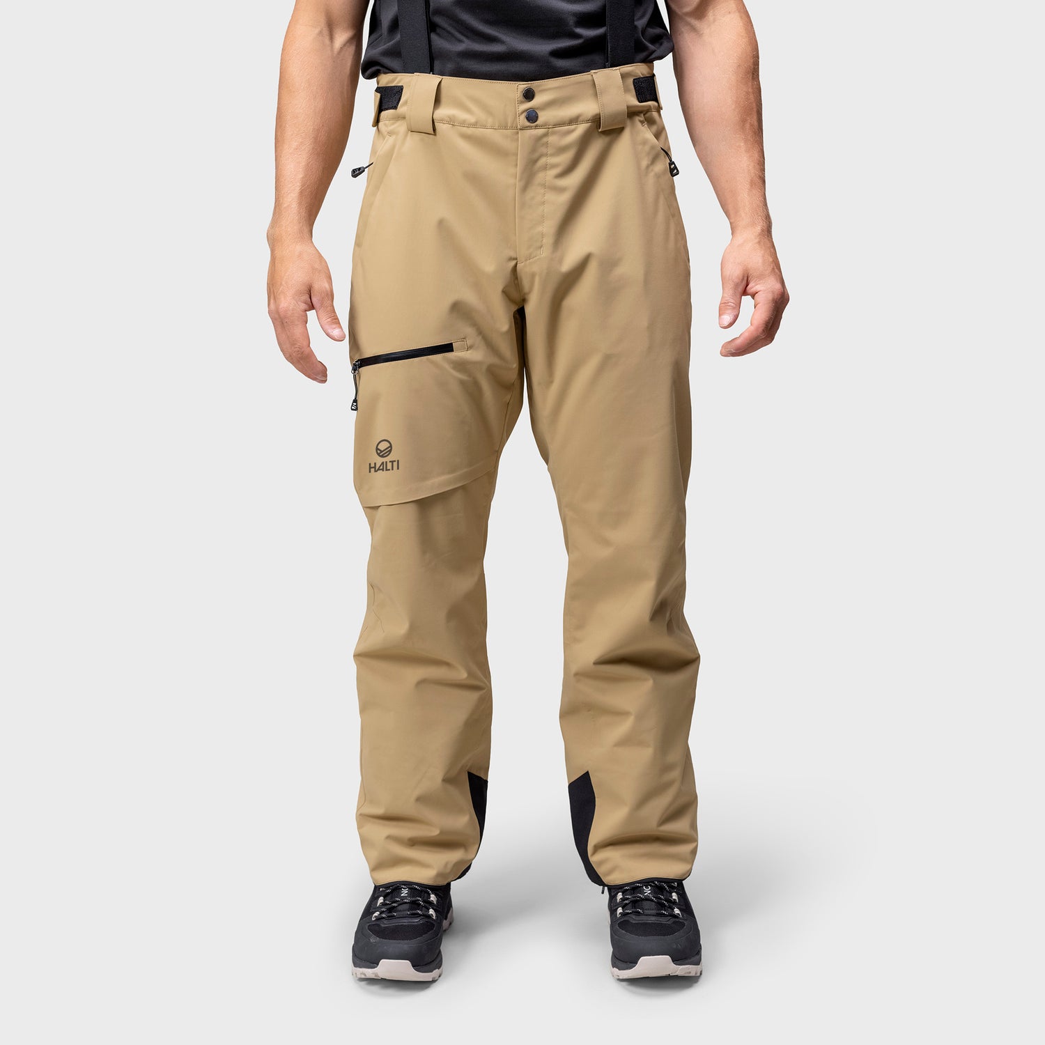 En person klädd i beige friluftsbyxor med hängslen, svarta skor och svart skjorta står mot en enfärgad bakgrund.