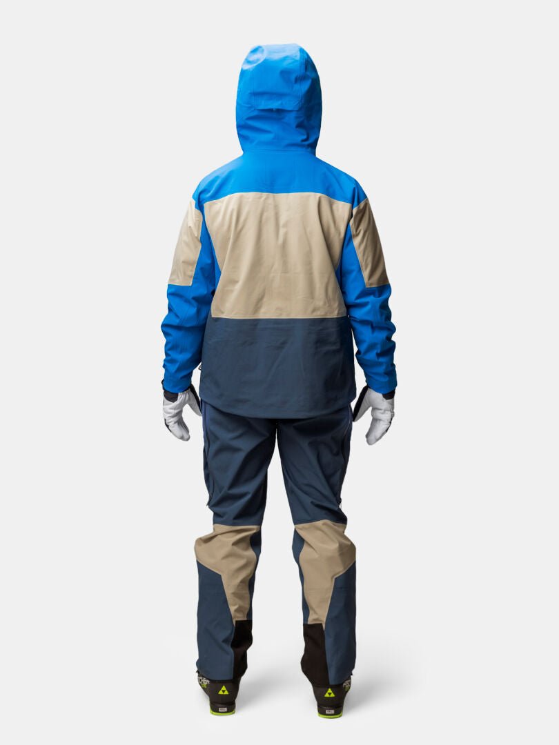 Bakifrånbild av en person som bär en blå och beige Halti Rizle Unisex 3L DX Skidjacka och matchande byxor samt vita handskar.