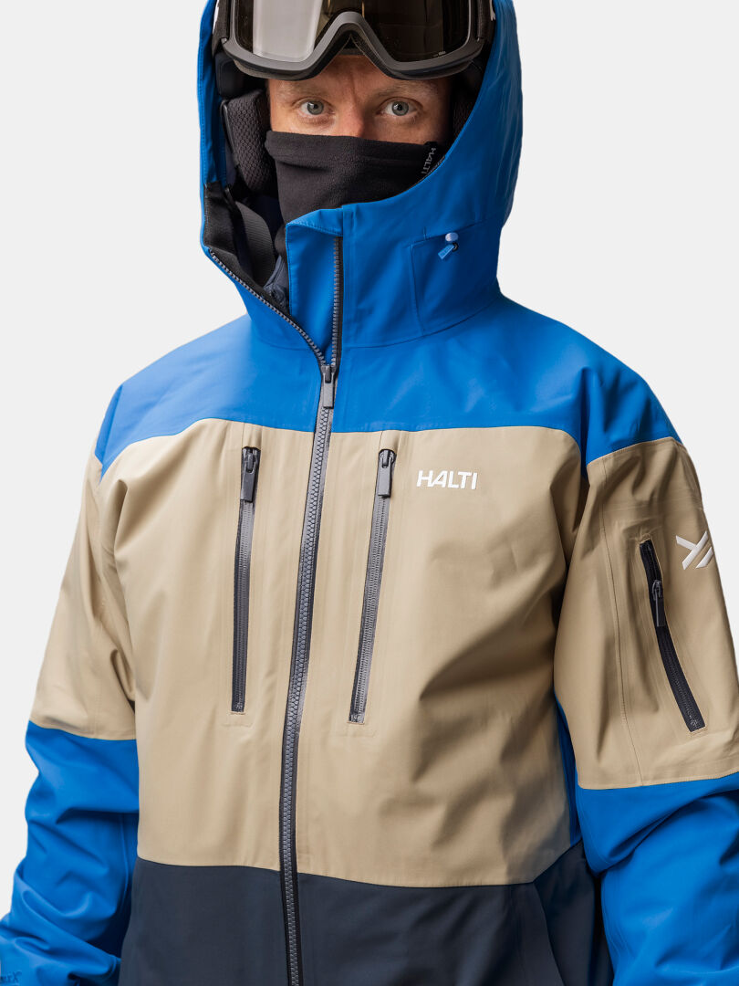 En person som bär en blå, beige och svart Halti Rizle Unisex 3L DX Skidjacka med huva, skidglasögon och ansiktsmask.