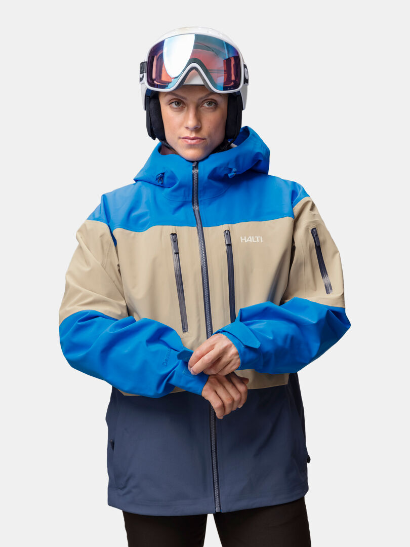 En person med skidhjälm och skidglasögon, klädd i en blå och beige Halti Rizle Unisex 3L DX Skidjacka.