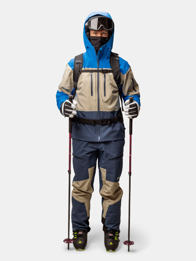 En person i blå och beige Halti Rizle 3L DX Skidjacka Unisex, skyddsglasögon, handskar och skidstavar står vänd framåt.