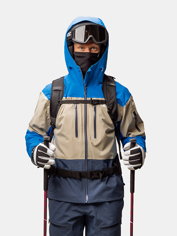 En skidåkare klädd i en blå och beige Halti Rizle Unisex 3L DX Skidjacka, hjälm, skidglasögon, ansiktsmask, ryggsäck och skidstavar.