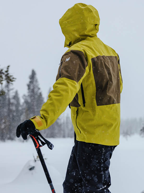 En person i gul och brun Halti Rizle 3L DX Skidjacka Unisex står i snön med en skidstav i handen, med ryggen mot kameran.