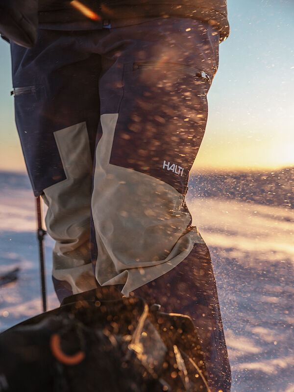 Närbild av en person i Halti Rizle 3L DX Skidbyxor Unisex, stående på snöig mark i solnedgång med snöblandat regn.