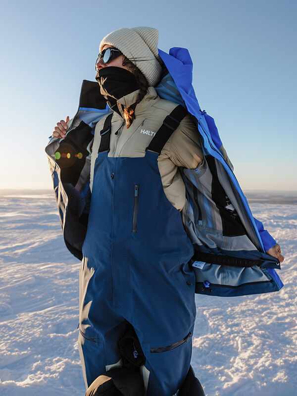 Person med solglasögon, mössa, mask och Halti Rizle 3L DX Skidbyxor Unisex opens en blågrå jacka på snöig mark.