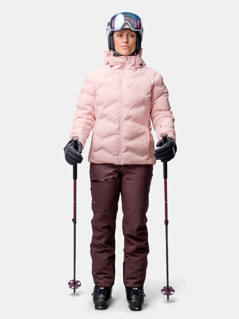 En person bär Halti Nordic Arcty II Skijacka Dam, bruna byxor, skidutrustning med hjälm, skidglasögon och stavar.