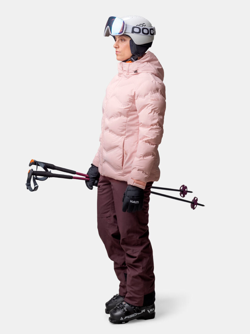 Person i skidutrustning håller i stavar, klädd i rosa Halti Nordic Arcty II Skijacka Dam och rödbruna byxor.