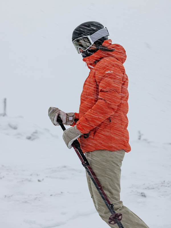 Person i Halti Nordic Arcty II Skijacka Dam och hjälm står i ett snöigt, blåsigt område och håller i skidstavarna.