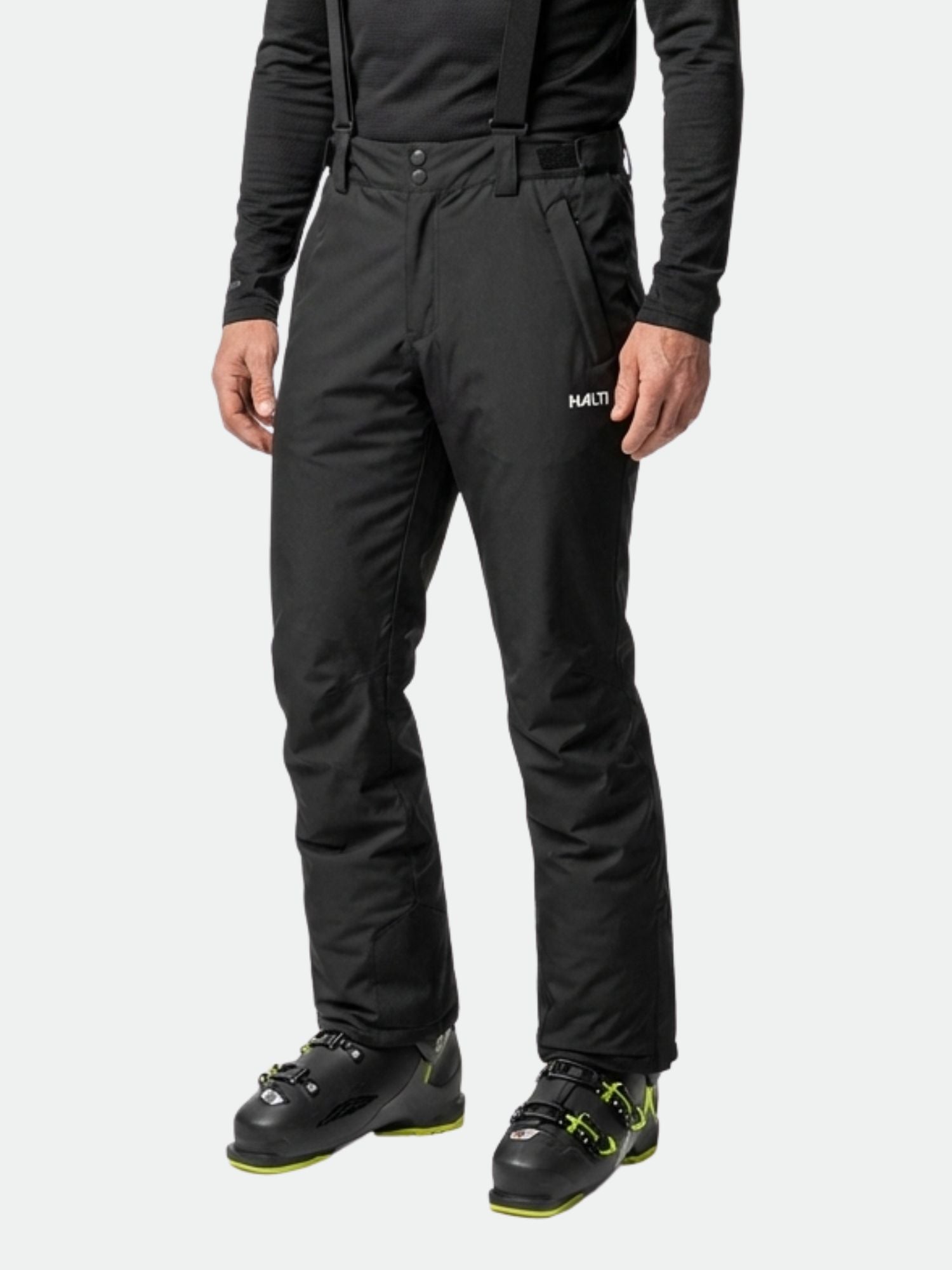 En person bär Halti Glades II DX skidbyxor, en svart topp och svarta pjäxor med gröna detaljer på en enfärgad bakgrund.