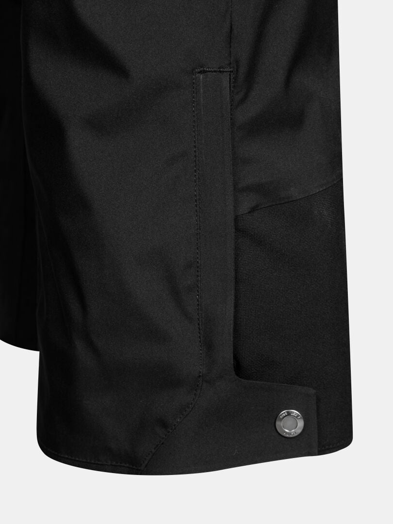 Närbild på ett byxben på ett par svarta Vertica II DX Skidbyxor för herrar, isolerade med PrimaLoft, med en sidficka och en metallknapp med tryckknapp i byxbenets nederkant.