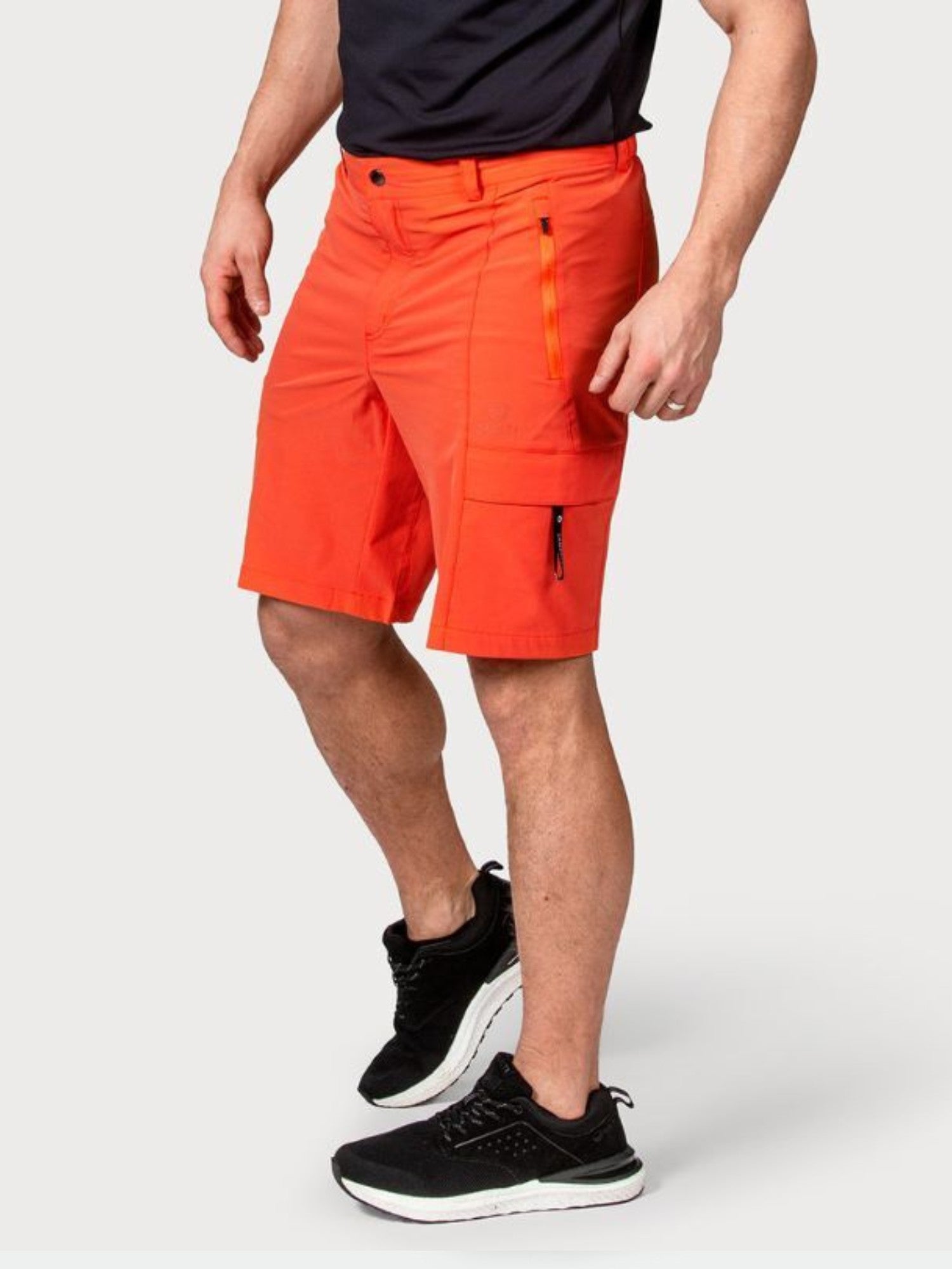 En man i Halti Reissu Herr Shorts, svart T-shirt och svarta sportskor står mot en enfärgad bakgrund.
