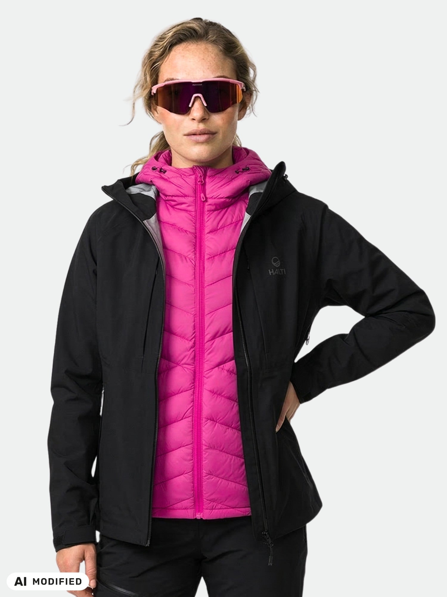 Modellen bär en Halti Kaarna Dam DrymaxX 3L-skaljacka över en rosa dunjacka och stora solglasögon, mot en ljus bakgrund.