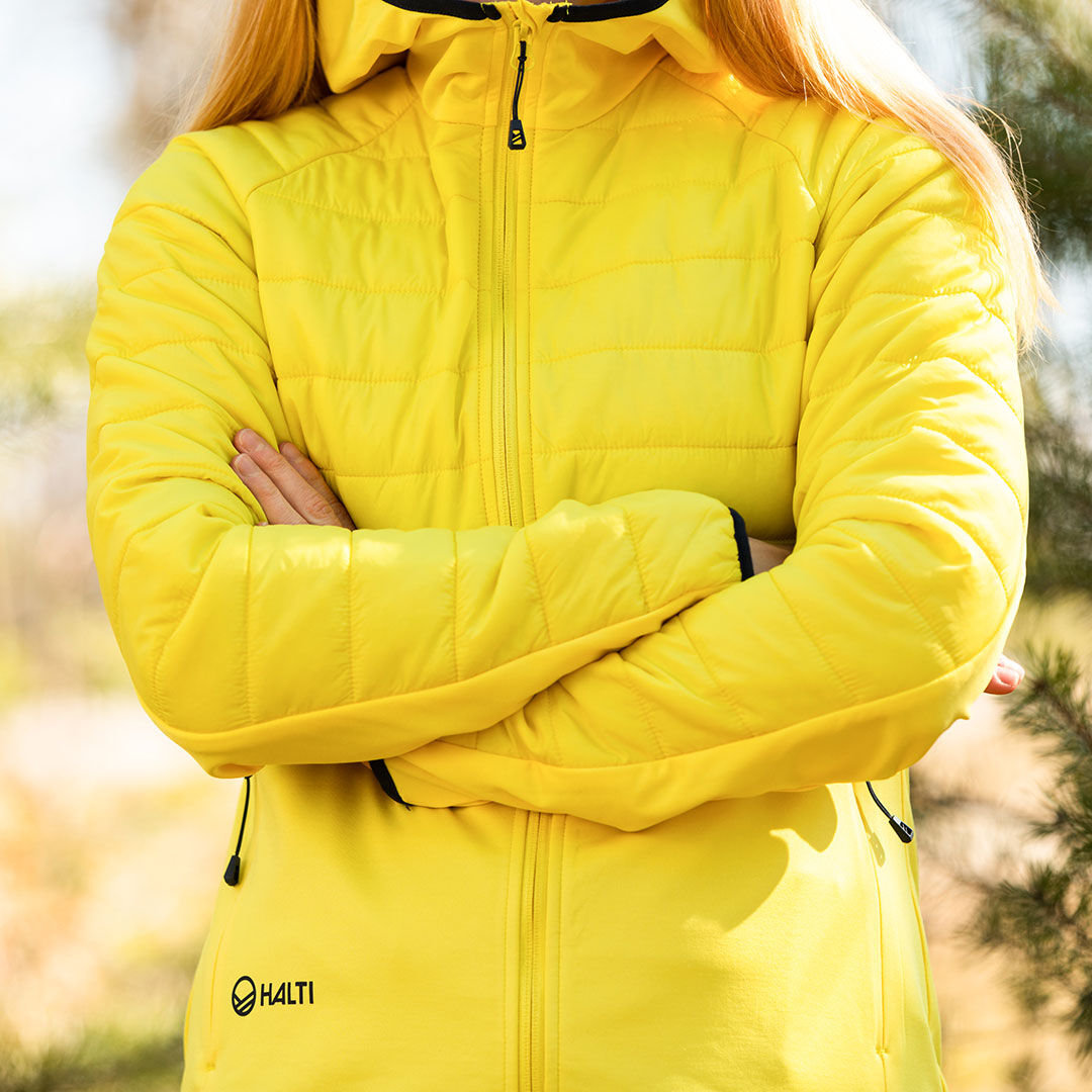 Person i Halti Veidnes Dam Mellanlager-jacka står utomhus med armarna i kors och njuter av friluftsliv.