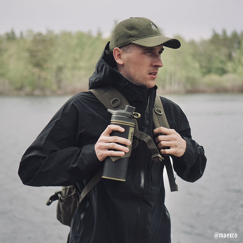 En man i Halti Kaarna Herr DrymaxX 3L Skaljacka och friluftsutrustning står vid en sjö, redo för vandring.
