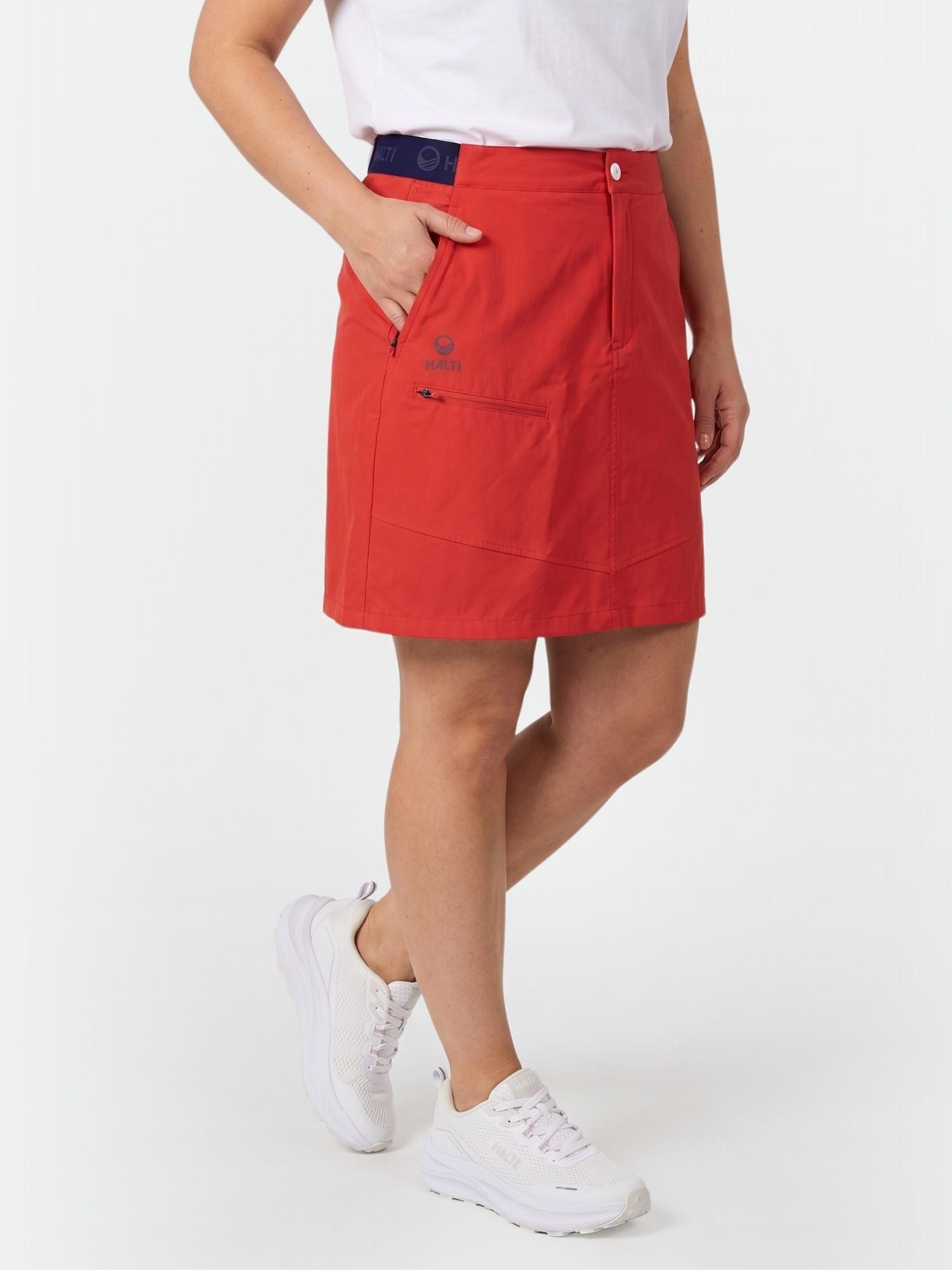 En person i röd Halti Pallas Dam Lite X-stretch Kjol, vit t-shirt och sneakers står mot en vit bakgrund.