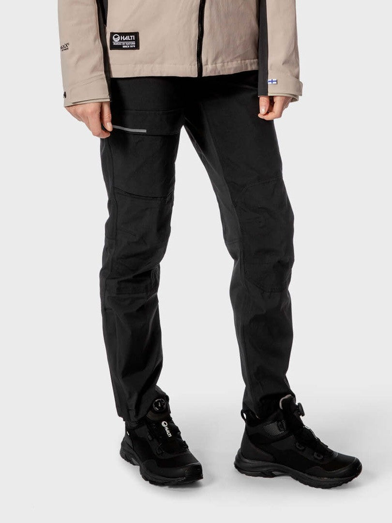 En person i beige jacka, Halti Hiker Dam DrymaxX Byxor och vandringskängor står mot en ljus bakgrund.