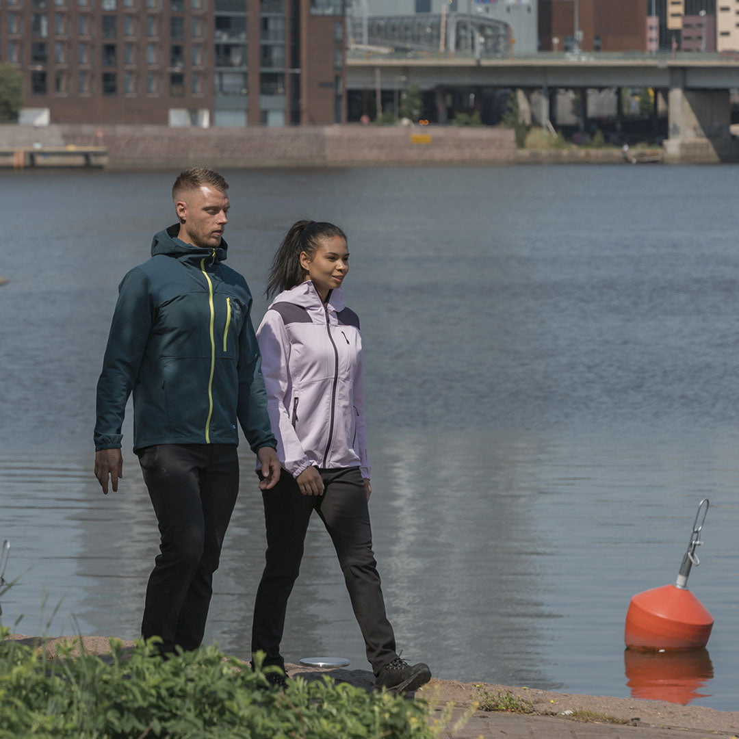 Två personer i jackor och Halti Pallas III Warm X-stretch Outdoor Byxor Dam promenerar vid en flodstrand med byggnader och en bro.