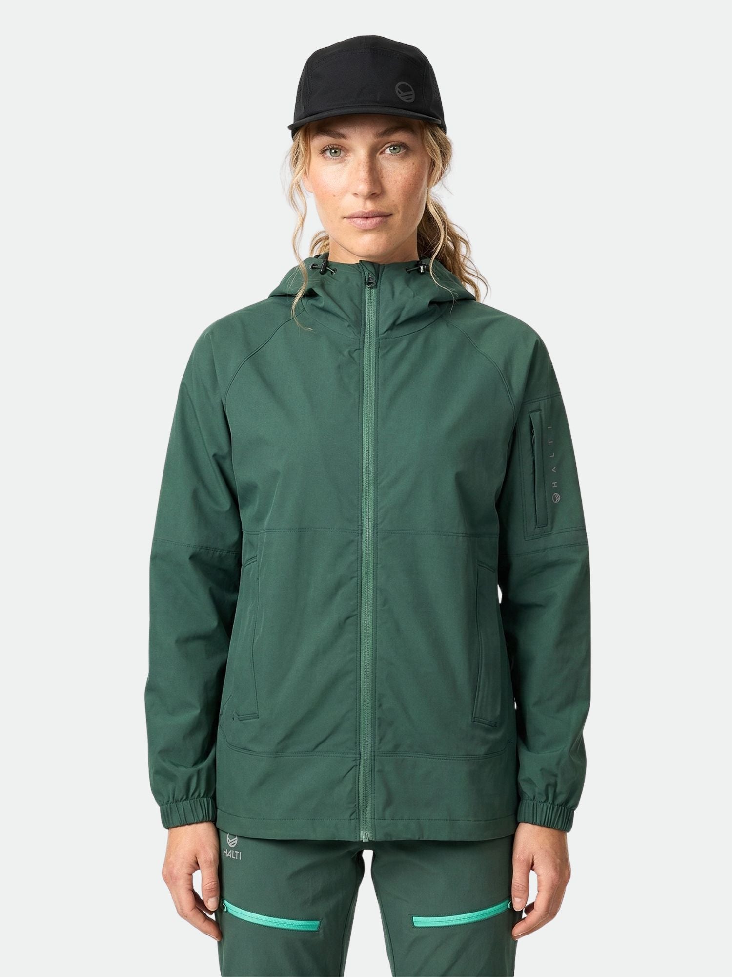 En person i mörkgrön Halti Pallas Jogger Lite Jacka Dam, matchande joggingbyxor och svart keps står mot en enfärgad bakgrund.