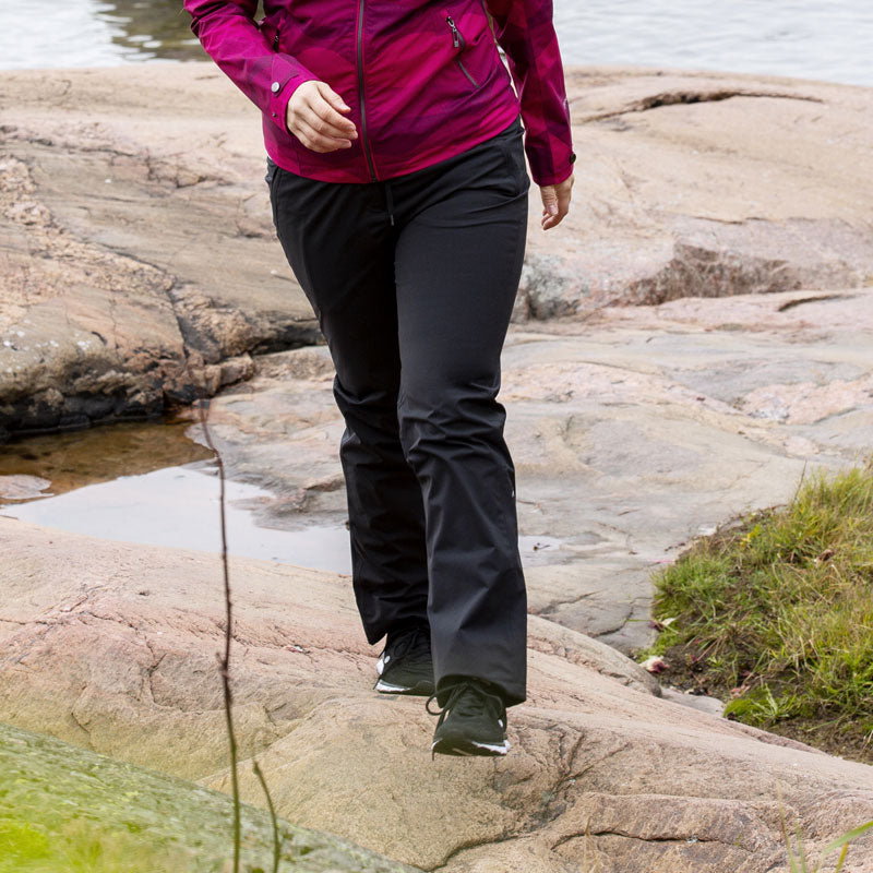En person i Halti Pine Plus Dam DrymaxX friluftsbyxor och en rödbrun jacka går i stenig terräng vid vattnet.