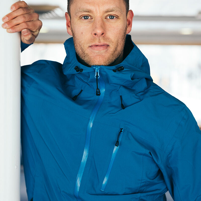 En man i en Halti Vattentät Skaljacka Herr står inomhus, vilar armen på en vit stång och tittar in i kameran.