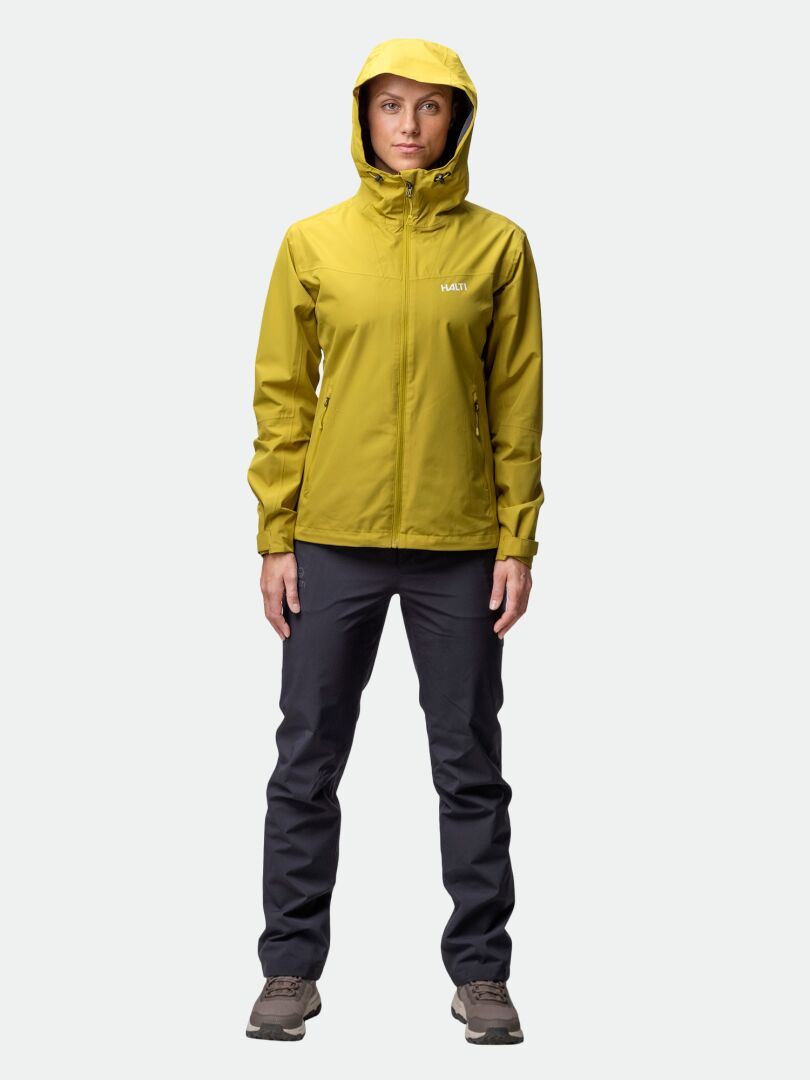 En person står framåtlutad i Halti Forter DX Skaljacka Dam, mörka byxor och bruna skor mot en enfärgad bakgrund.