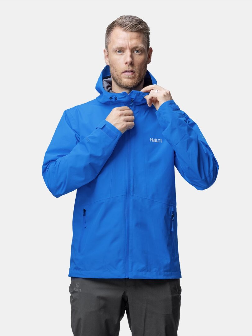 En man drar upp dragkedjan på Halti Forter DX Skaljacka Herr, vänd mot kameran mot en vit bakgrund.