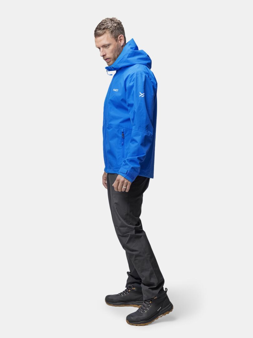 En man i blå Halti Forter DX Skaljacka, svarta byxor och vandringskängor står mot en vit bakgrund.