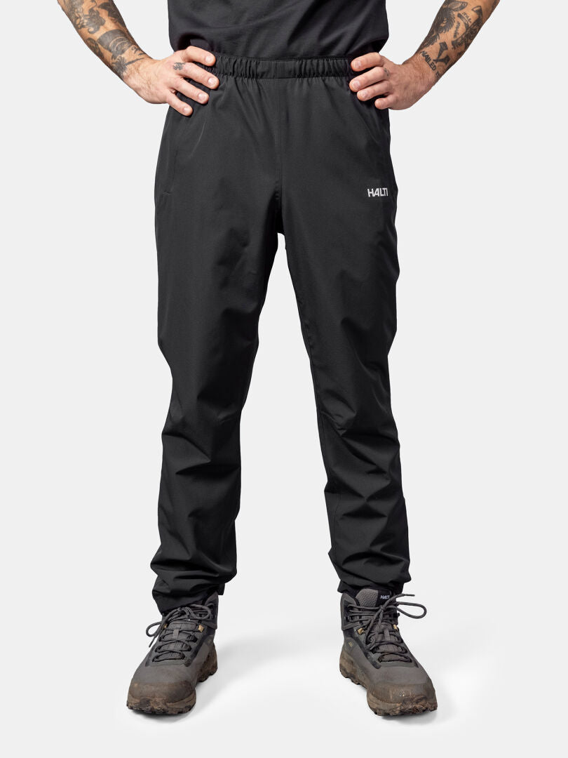 En person i Halti Forter DX Skalbyxor Herr, svart skjorta och leriga stövlar står mot en ljus bakgrund.