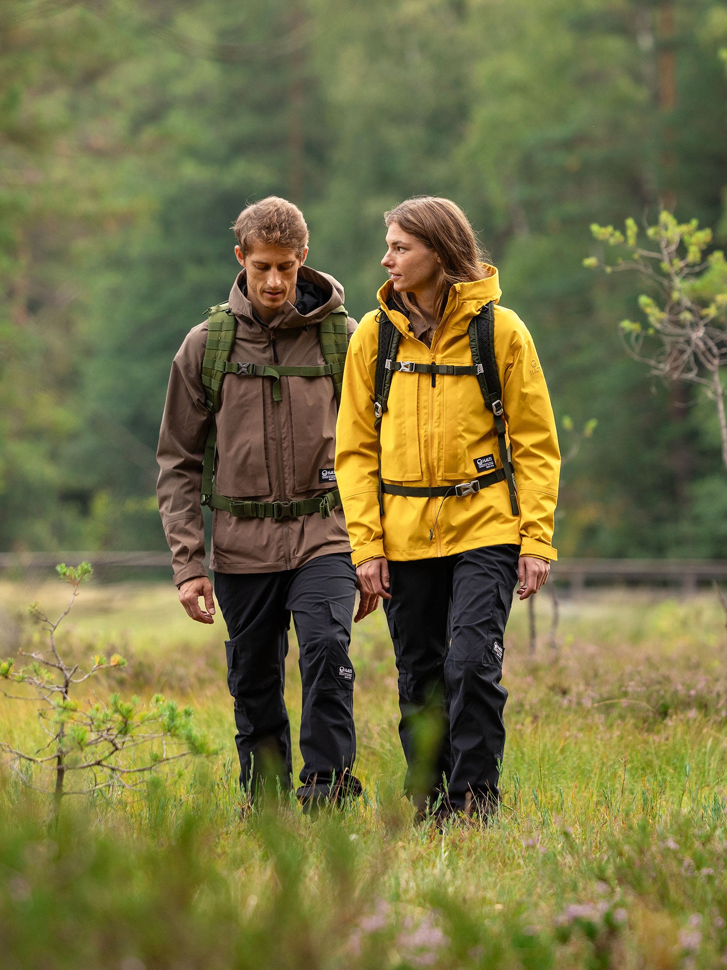 Två personer i Halti Hiker II DrymaxX Skaljacka-dammen går tillsammans genom gräset, redo för vandring bland träden.