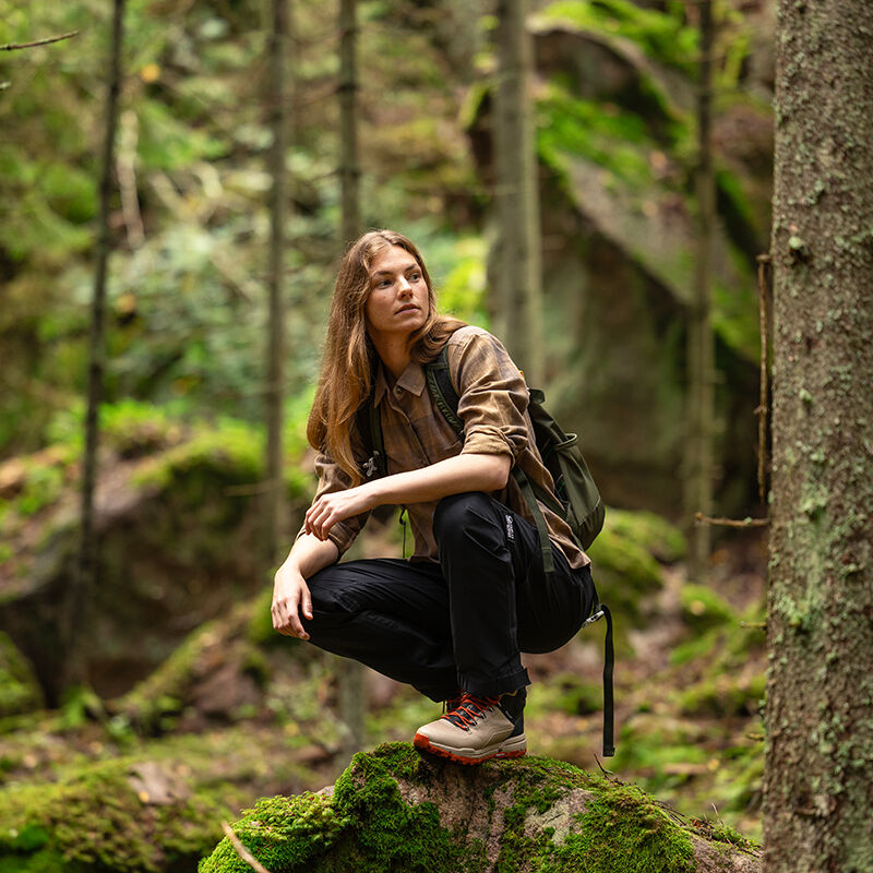 Iklädd Halti Hiker II DrymaxX Skalbyxor Dam sitter hon på huk på en mossig sten, redo att vandra i skogen med sin ryggsäck.