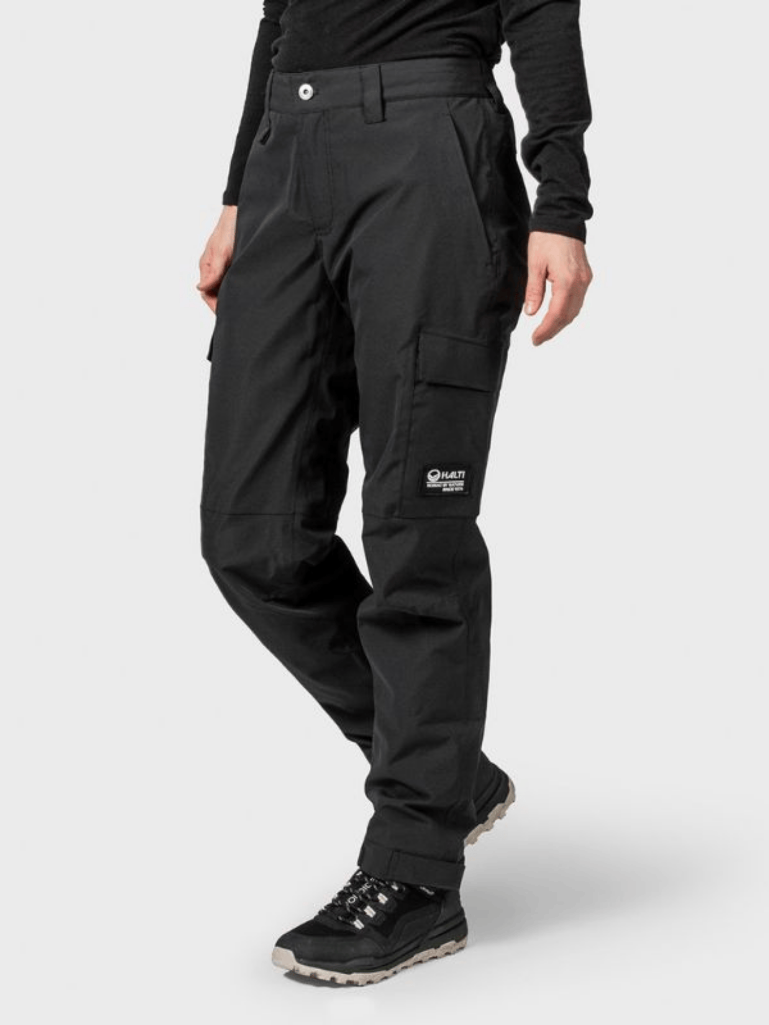 En person i Halti Hiker II DrymaxX skalbyxor för dam, svart långärmad topp och vandringskor står mot enkel bakgrund.