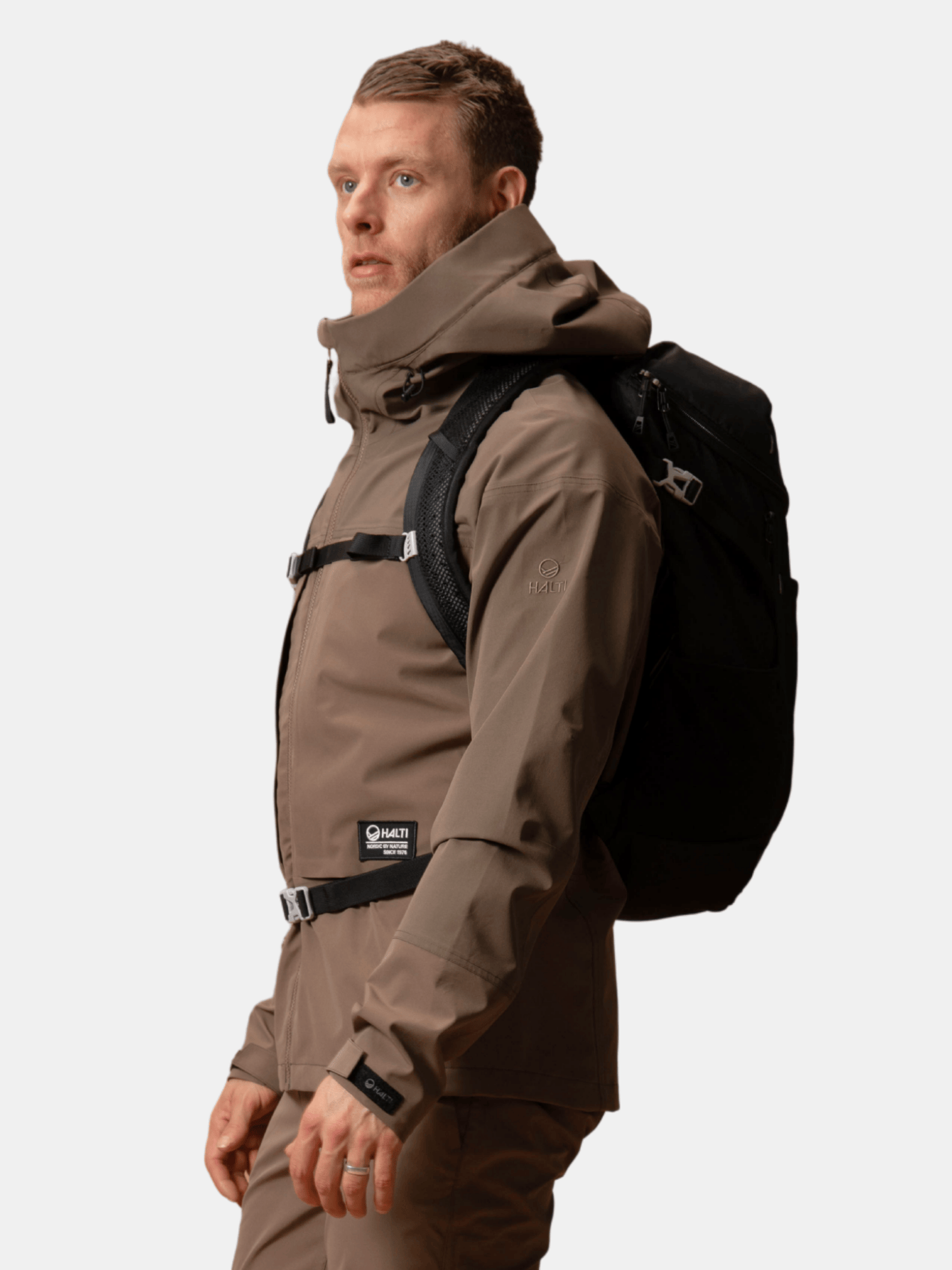 En person i Halti Hiker II DrymaxX Skaljacka Herr och en svart ryggsäck står mot en ljus bakgrund.