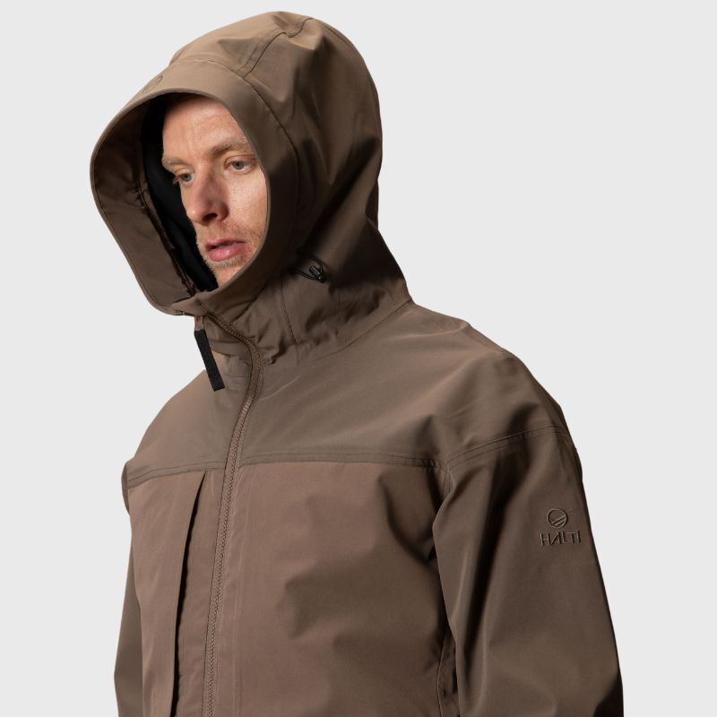 En man i en brun Halti Hiker II DX Skaljacka Herr med dragkedja tittar ner mot en ljus bakgrund.